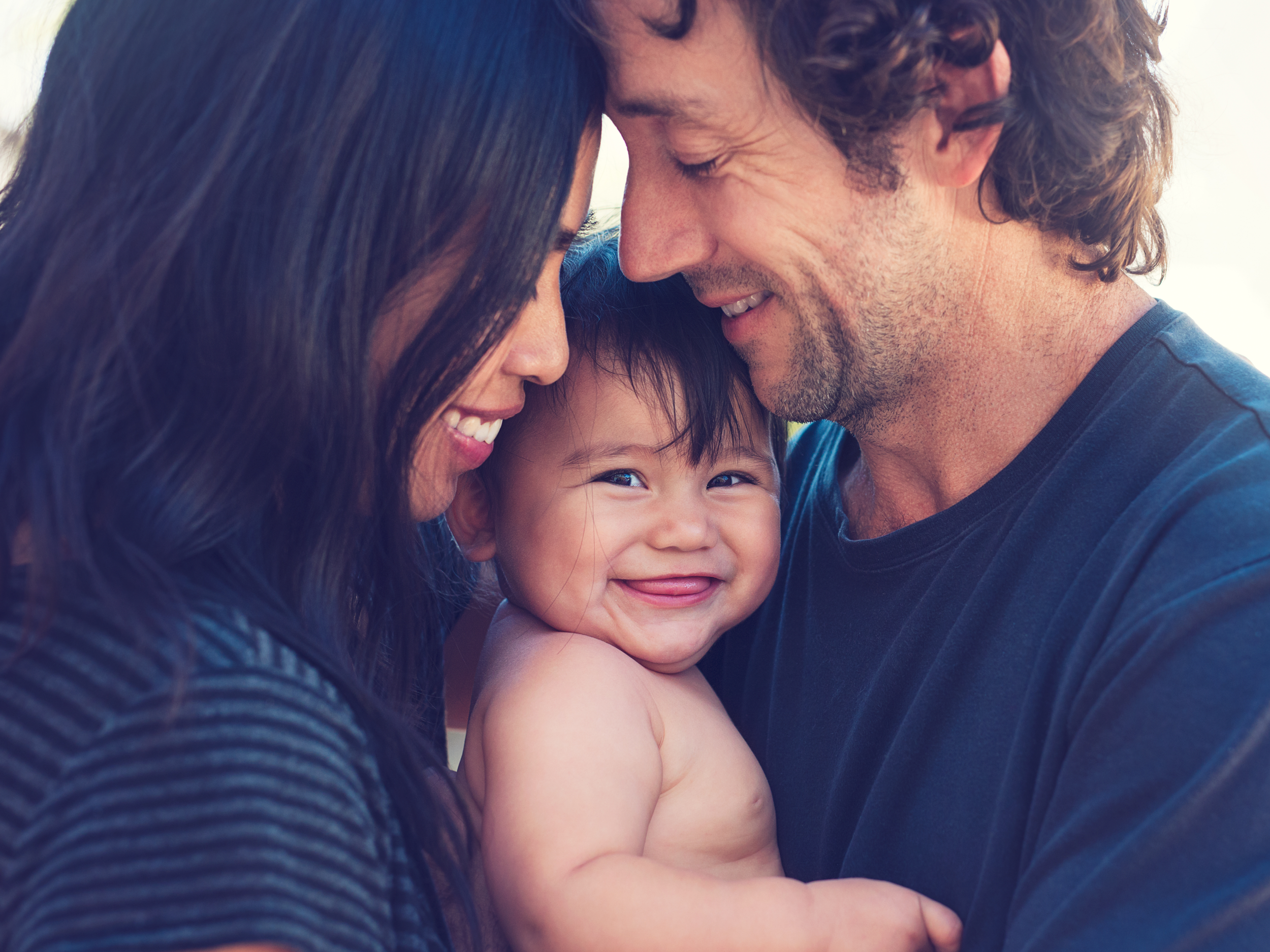 Three people, a man, a woman and a baby in a close hug. Baby is smiling at the camera.