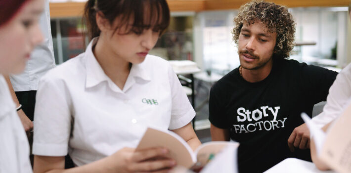 Five people in a library where a young person is sharing something in her book with a man in a black t-shirt who is looking at the book.