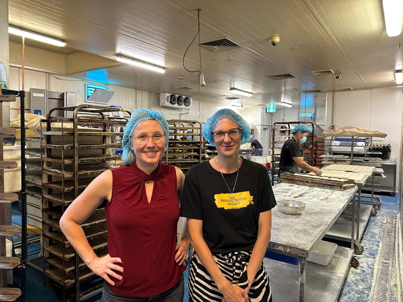 Two women in a commercial kitchen, wearing hairnets and smiling at the camera.