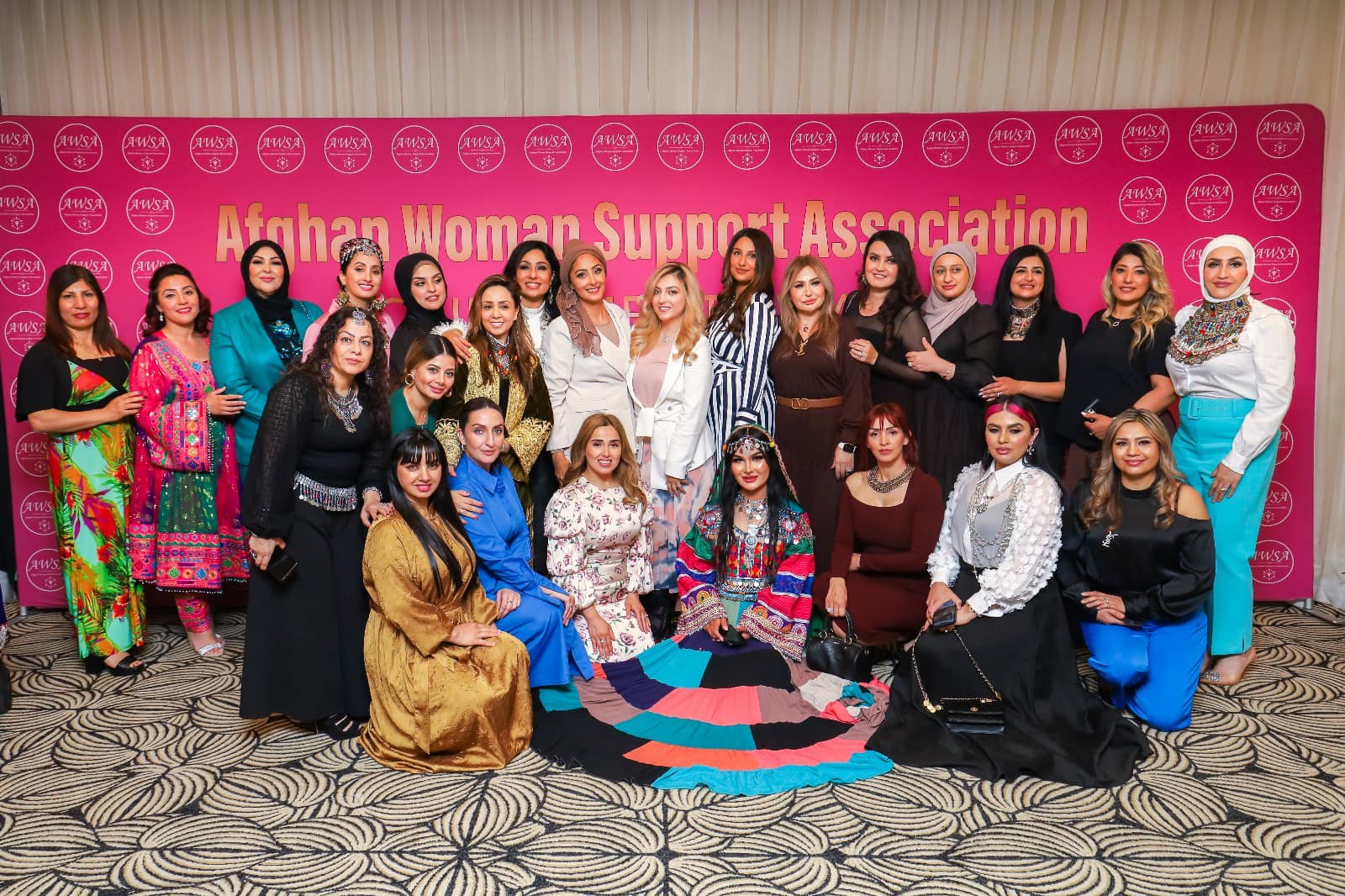 25 women standing in front of a pink media wall with the Afghan Women Support Association signage.